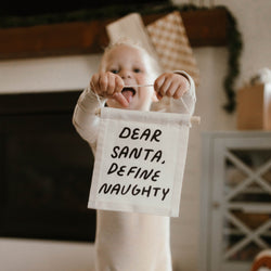 Child holding a humorous holiday sign reading "Dear Santa, Define Naughty" in a festive living room setting.