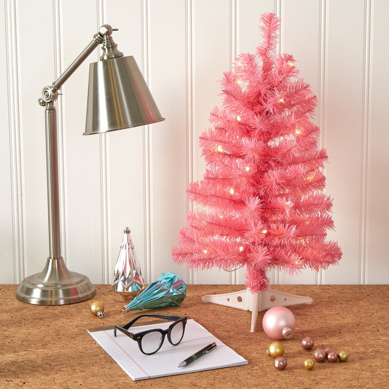 Small pink tabletop Christmas tree with warm string lights on a cork desk beside a metal task lamp, holiday ornaments, eyeglasses and notepad