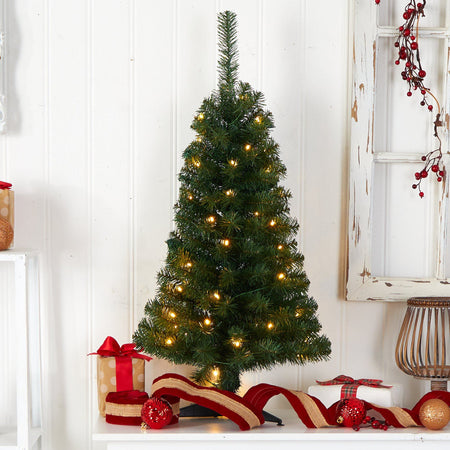 Pre-lit artificial tabletop Christmas tree with warm white LED lights, red ribbon, ornaments and wrapped gifts against a rustic white shiplap backdrop