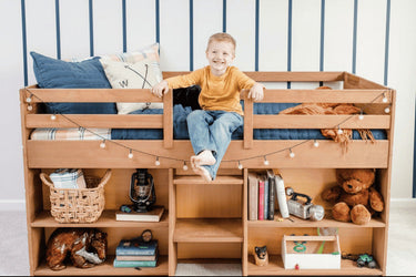 Child sitting on a wooden low loft bed with books and toys in a room with striped walls.