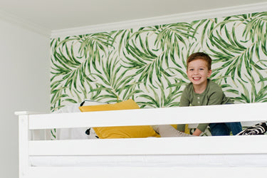 Child peeking over a white railing with green leaf-patterned wallpaper in the background.