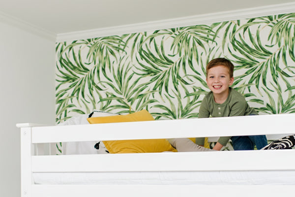 Child peeking over a white railing with green leaf-patterned wallpaper in the background.