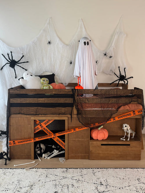A Halloween-themed wooden storage cabinet decorated with black tarps, orange warning tape, spider cutouts, a ghost figure, pumpkins, and skeleton props, set against a beige wall and white web backdrop.