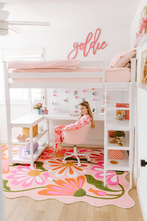 Pink-themed room with a girl sitting at a desk, floral rug, and 'goldie' sign.