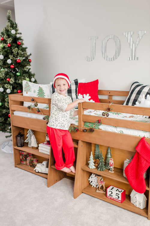 Smiling child in red pajamas and knit Santa hat climbing a wooden bunk bed in a cozy Christmas bedroom with decorated tree, JOY wall letters, holiday garland and countdown blocks.
