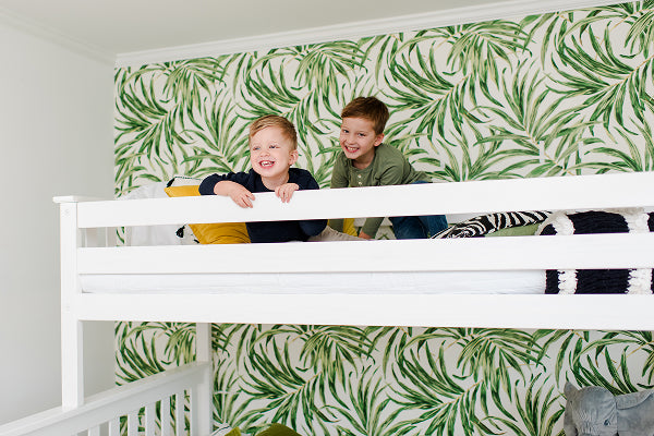 Two children smiling on a white bunk bed in a bright kids' room with green tropical leaf wallpaper and playful modern decor.