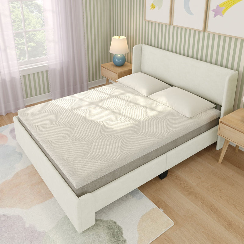 Upholstered platform bed with textured cream mattress and two pillows in a light green striped bedroom with wooden nightstands, blue lamp, pastel watercolor rug and sunlight from window