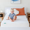Smiling toddler sitting on a white gel memory foam mattress in a bright modern bedroom with an orange pillow and nightstand