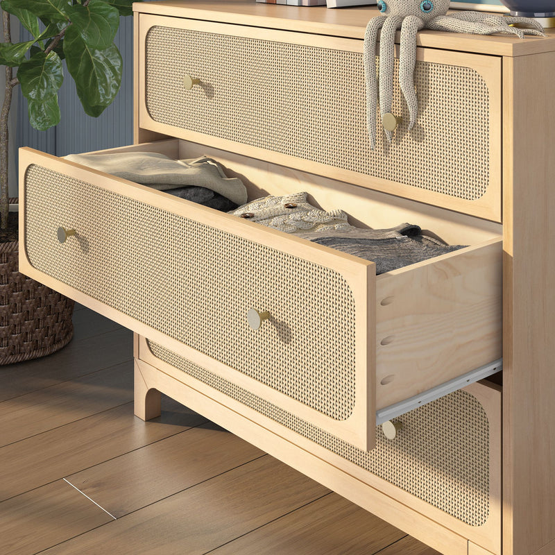 Light wood dresser with cane-woven rattan drawer fronts and brass knobs, open drawer revealing folded sweaters and linens in a modern boho bedroom setting with hardwood floors and potted plant