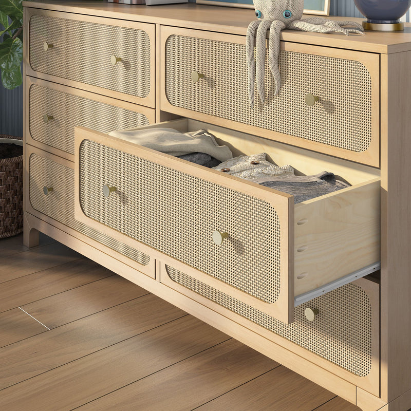 Woven cane-front natural wood dresser with brass knobs and open drawer containing folded clothes on hardwood floor — boho nursery or bedroom storage solution