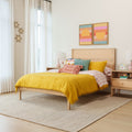Bright modern bedroom with yellow comforter, wooden headboard and bed frame, colorful throw pillows, striped sheets, abstract sun wall art, sheer curtains and bedside lamp