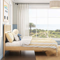 Sunlit coastal bedroom with natural wood bed, yellow-striped bedding and plush toy, sheer curtains framing a large window with palm tree and ocean view.