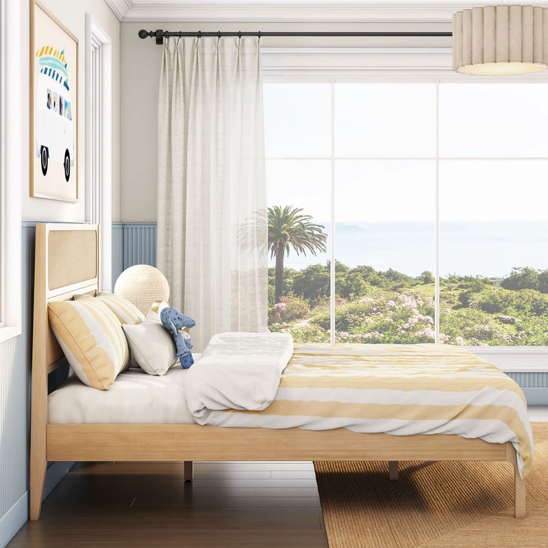 Sunlit coastal bedroom with natural wood bed, yellow-striped bedding and plush toy, sheer curtains framing a large window with palm tree and ocean view.