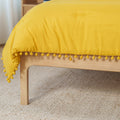 Close-up of mustard yellow pom-pom duvet draped over a light wood bed frame on a textured natural woven rug