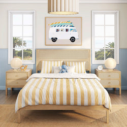 Coastal kids' bedroom with yellow-striped bedding, rattan headboard, wood nightstands, surfboard-van wall art and ocean-view windows.