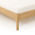 Close-up of a white quilted mattress cover on a light wood platform bed frame corner, showing mattress texture and bed frame detail