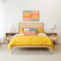 Modern bedroom with bright yellow duvet, textured throw pillows, woven rattan headboard, wooden nightstands with lamps and colorful framed wall art