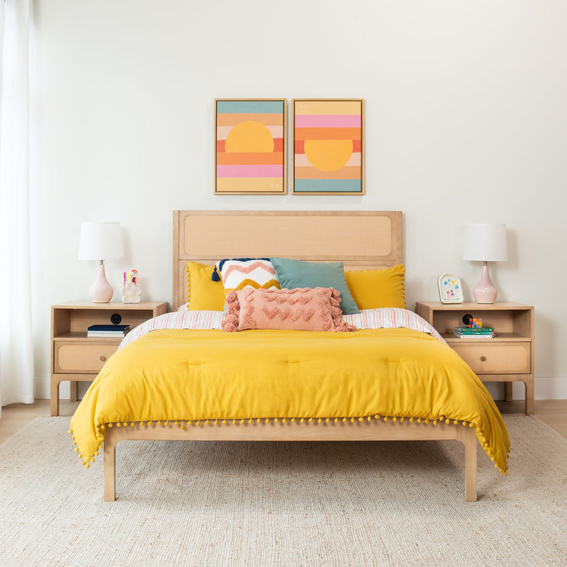 Modern bedroom with bright yellow duvet, textured throw pillows, woven rattan headboard, wooden nightstands with lamps and colorful framed wall art