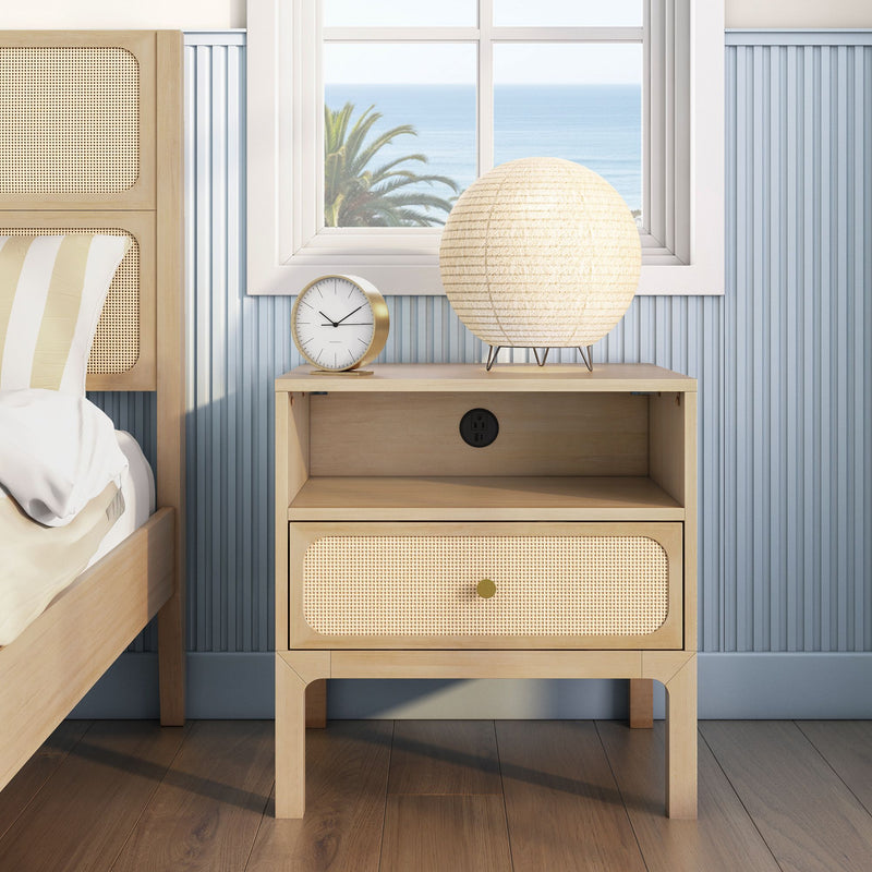 Rattan nightstand in coastal bedroom — natural wood bedside table with woven drawer, round paper-lantern lamp and gold-accent clock on blue beadboard wall with ocean view window and palm tree