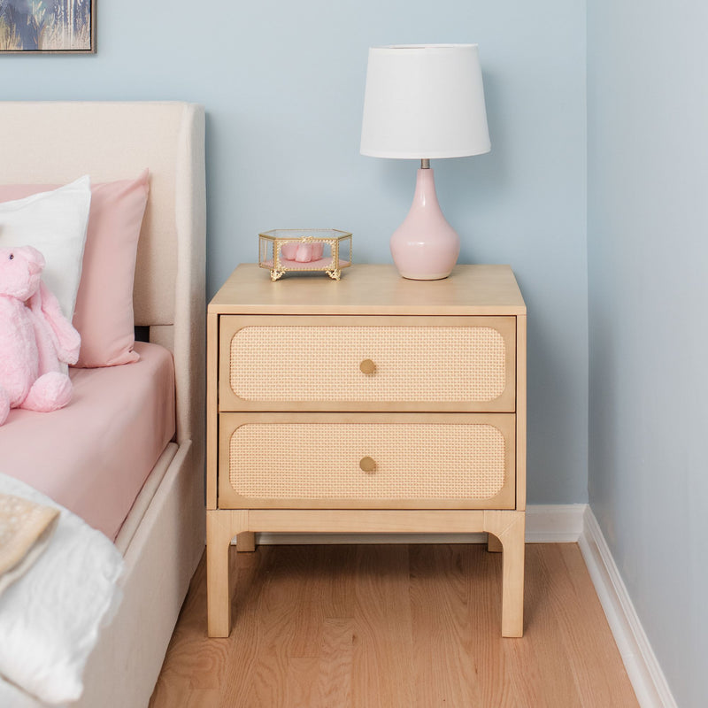 Light wood rattan two-drawer nightstand with pink ceramic lamp and glass jewelry box beside pastel pink bed in modern bedroom