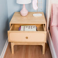 Light wood mid-century nightstand with open drawer showing "The Book With No Pictures", pink ceramic lamp, gold jewelry stand and pastel pink bedding on hardwood floor