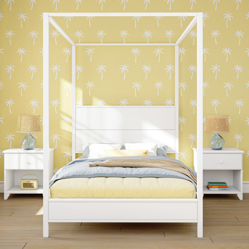 White canopy bed with yellow palm leaf wallpaper, wooden floor, and bedside tables with lamps.