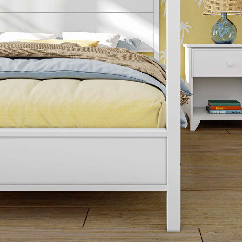 White wooden bed with yellow duvet and blue throw in coastal bedroom; white nightstand with lamp and books on hardwood floor.