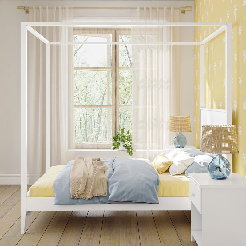 Sunlit modern coastal bedroom with white four-poster canopy bed, blue and yellow bedding, palm-print yellow wallpaper, sheer curtains, wooden floor, bedside tables with blue glass lamps and a potted plant on the windowsill