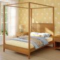 Wooden four-poster canopy bed in sunny bedroom with yellow palm-print wallpaper, natural wood nightstand, lamp and layered bedding