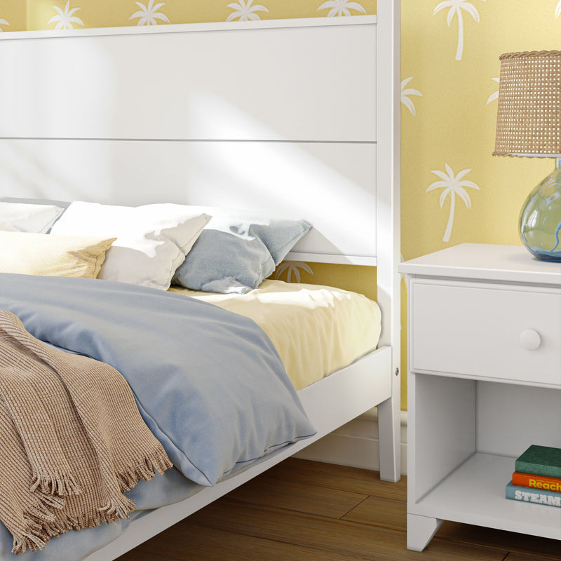 Cozy bedroom with white bed frame, colorful pillows, and a stylish nightstand against palm-printed wallpaper.