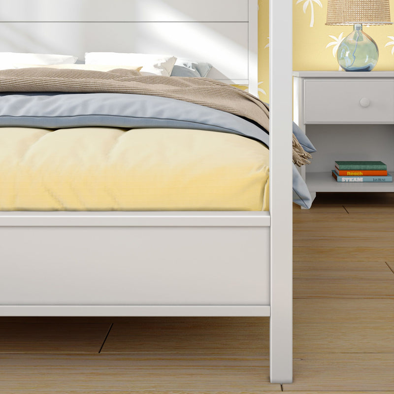 Close-up of a white four-poster bed with yellow duvet, blue throw and beige blanket in a modern bedroom with wooden floor, bedside table, glass lamp, and palm-print wallpaper