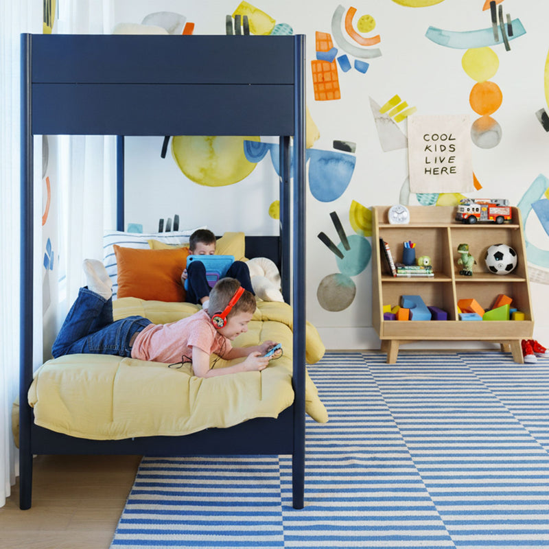 Modern kids bedroom with navy bunk bed and yellow bedding, colorful abstract wall mural, wooden toy shelf, striped blue rug, two children playing with a tablet and handheld game