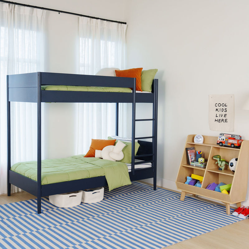 Bright modern kids' bedroom featuring a navy bunk bed with ladder, green bedding and orange pillows, blue-striped rug and wooden toy organizer with children's toys and storage