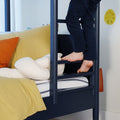 Child climbing navy blue bunk bed ladder barefoot beside yellow bedding and white plush toy in a modern kids bedroom