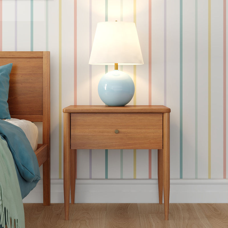 Bedroom scene: mid-century modern wooden nightstand with single drawer and blue ceramic bedside lamp on hardwood floor, pastel striped wallpaper and blue bedding
