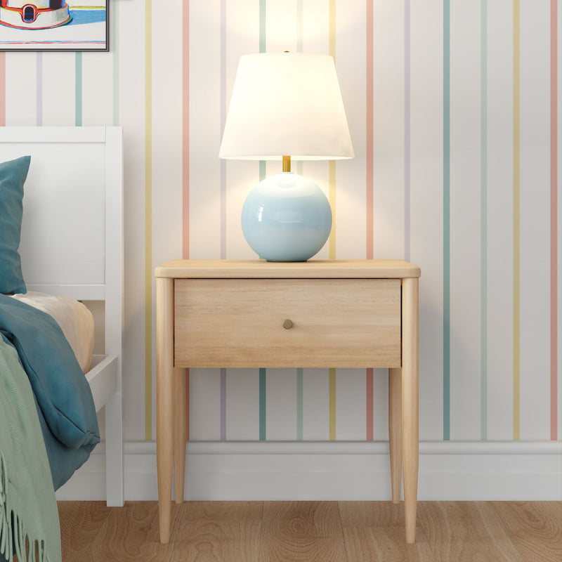 Modern Scandinavian wooden nightstand with blue ceramic table lamp on pastel striped wallpaper beside white bed with teal bedding