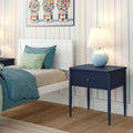 Navy blue bedside table with round blue lamp next to a white bed with teal and mint bedding, pastel striped wallpaper and wood floor