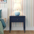 Navy blue nightstand with single drawer and round blue ceramic lamp on top against pastel vertical-striped wallpaper with white bed frame and teal bedding in a modern bedroom