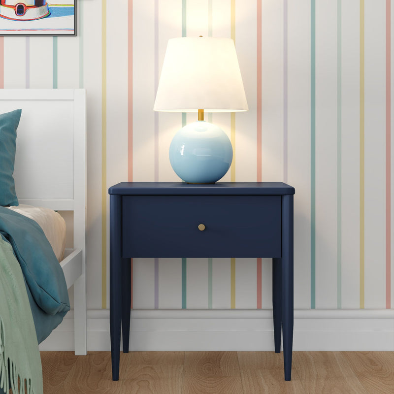 Navy blue nightstand with single drawer and round blue ceramic lamp on top against pastel vertical-striped wallpaper with white bed frame and teal bedding in a modern bedroom