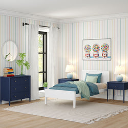 Contemporary children's bedroom with white twin bed, teal bedding and throw, navy blue nightstands and dresser, pastel vertical striped wallpaper, colorful gumball machine wall art, white curtains over black-trim window, light wood floors and neutral area rug