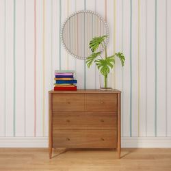 Mid-century wooden dresser with colorful stacked books, glass vase with monstera leaves, and round beaded mirror on pastel striped wallpaper