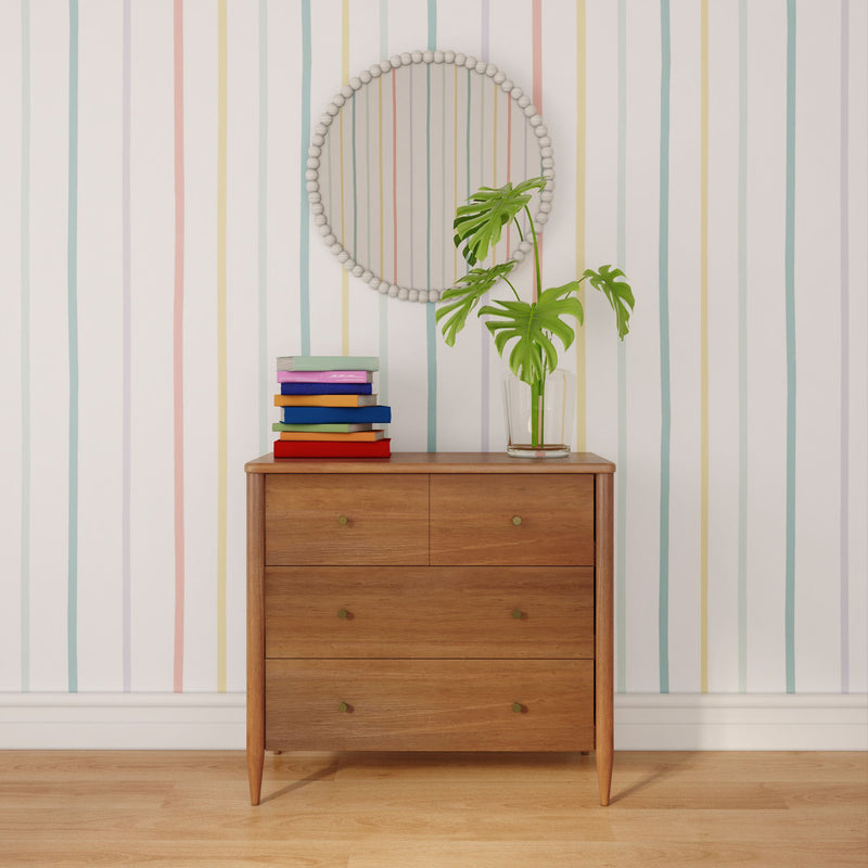 Mid-century wooden dresser with colorful stacked books, glass vase with monstera leaves, and round beaded mirror on pastel striped wallpaper