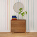 Mid-century modern wooden dresser styled with stacked colorful books, glass vase with monstera leaves, round beaded mirror, and pastel striped wallpaper on a hardwood floor