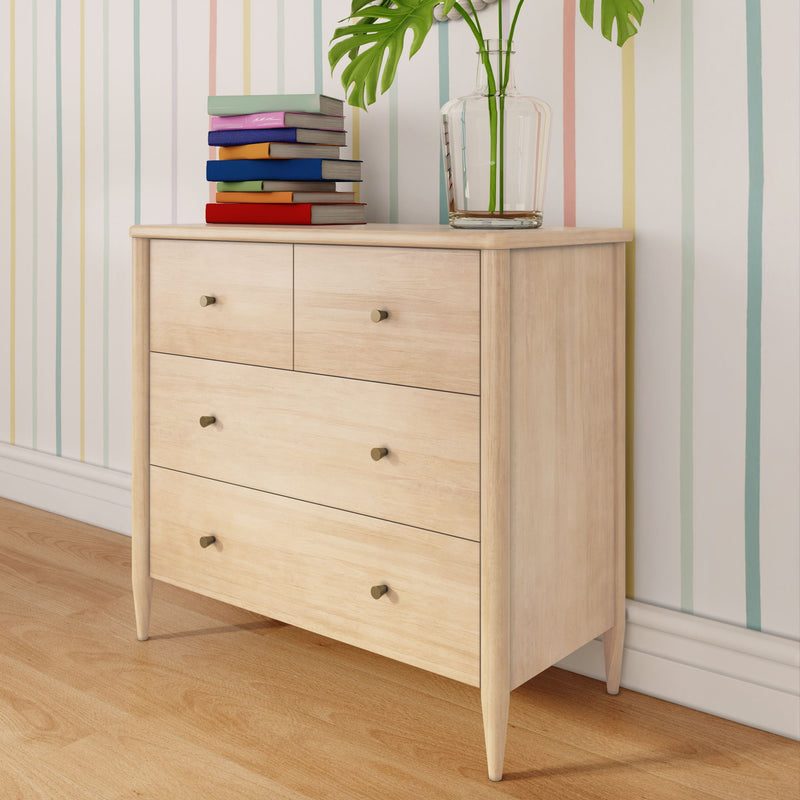 Light wood mid-century modern dresser with three drawers on hardwood floor, stacked colorful books and a glass vase with green leaves against pastel striped wallpaper
