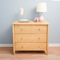 Light wood three-drawer dresser styled with a pink ceramic table lamp, gold jewelry stand, stacked books and a pink instant camera against a pale blue wall on hardwood floor