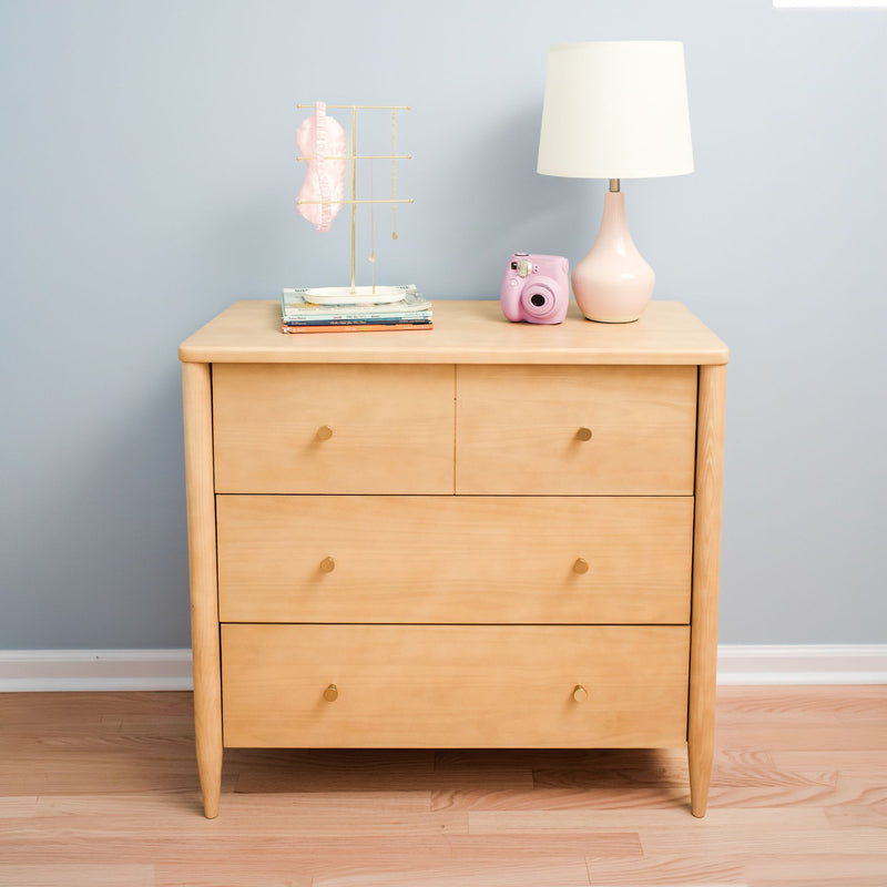 Light wood three-drawer dresser styled with a pink ceramic table lamp, gold jewelry stand, stacked books and a pink instant camera against a pale blue wall on hardwood floor