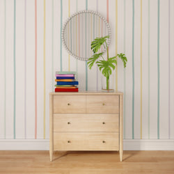 Light wood dresser with drawers, round beaded wall mirror, stacked colorful books and glass vase with monstera plant against pastel vertical striped wallpaper
