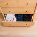 Open light-wood dresser drawer with neatly folded jeans, shirts and black garments on hardwood bedroom floor