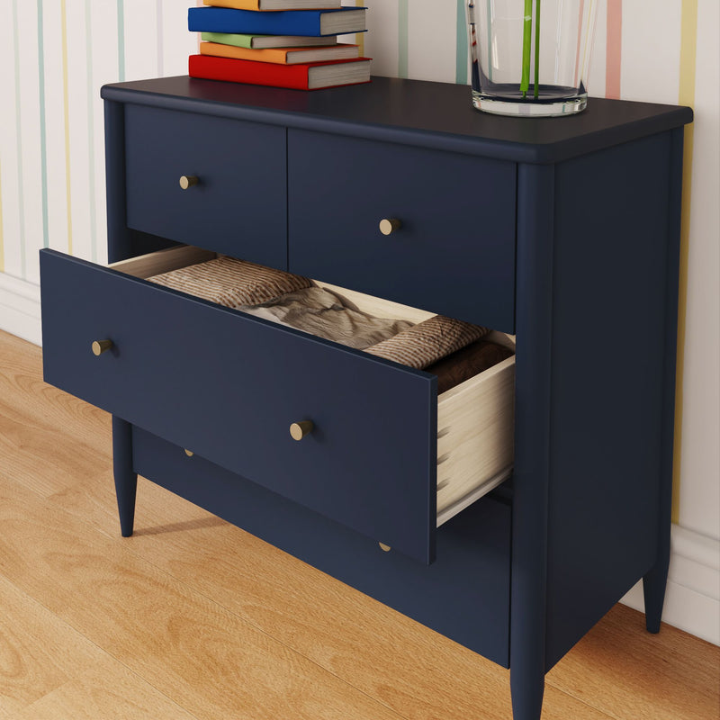 Navy blue mid-century modern dresser with brass knobs and an open drawer of folded linens, books and a glass vase on top against a pastel striped wallpaper on hardwood floor