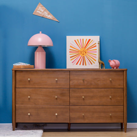 Mid-century modern six-drawer wooden dresser against blue wall with pink mushroom lamp, colorful sun art print, brass giraffe figurine and pink piggy bank, children's room decor